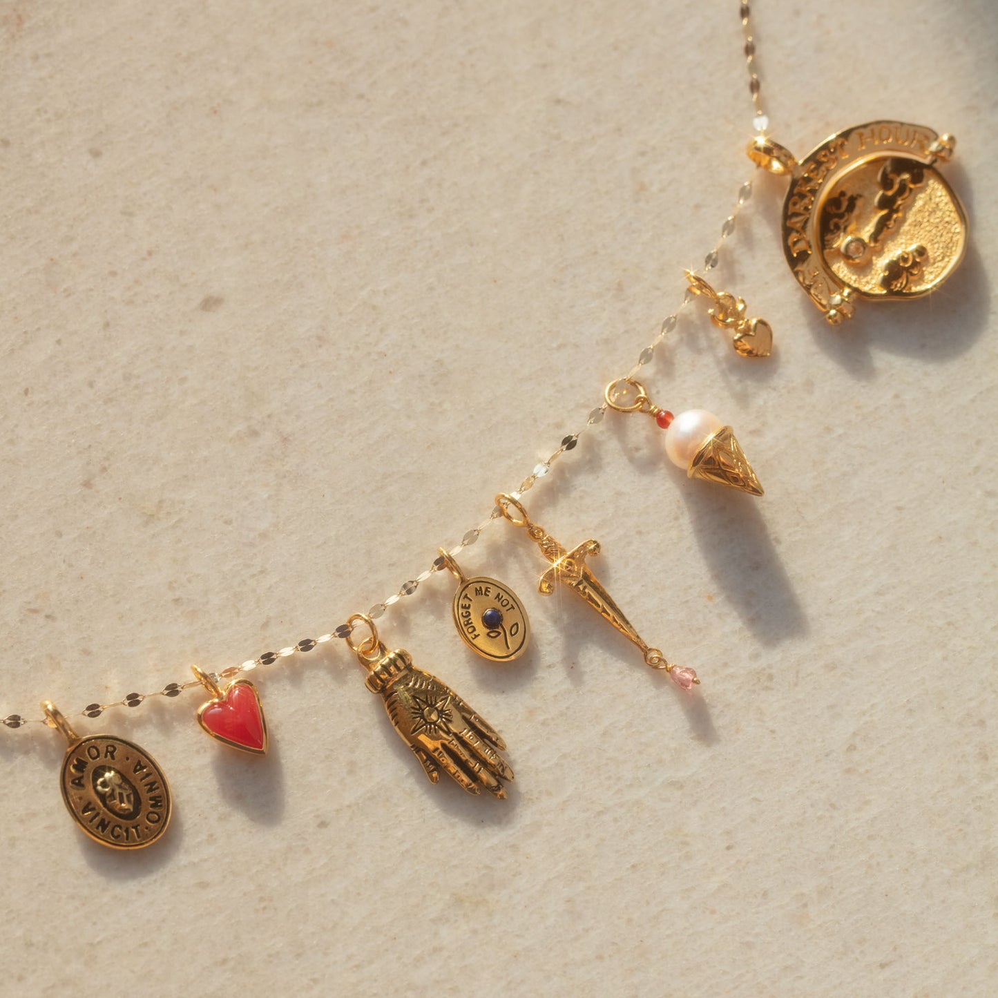 The Charms by Hunt of Hounds necklace features gold vermeil and assorted pendants—such as a red heart, hand, umbrella, pearl-topped ice cream cone, coins, and other charms—displayed on a light textured surface.