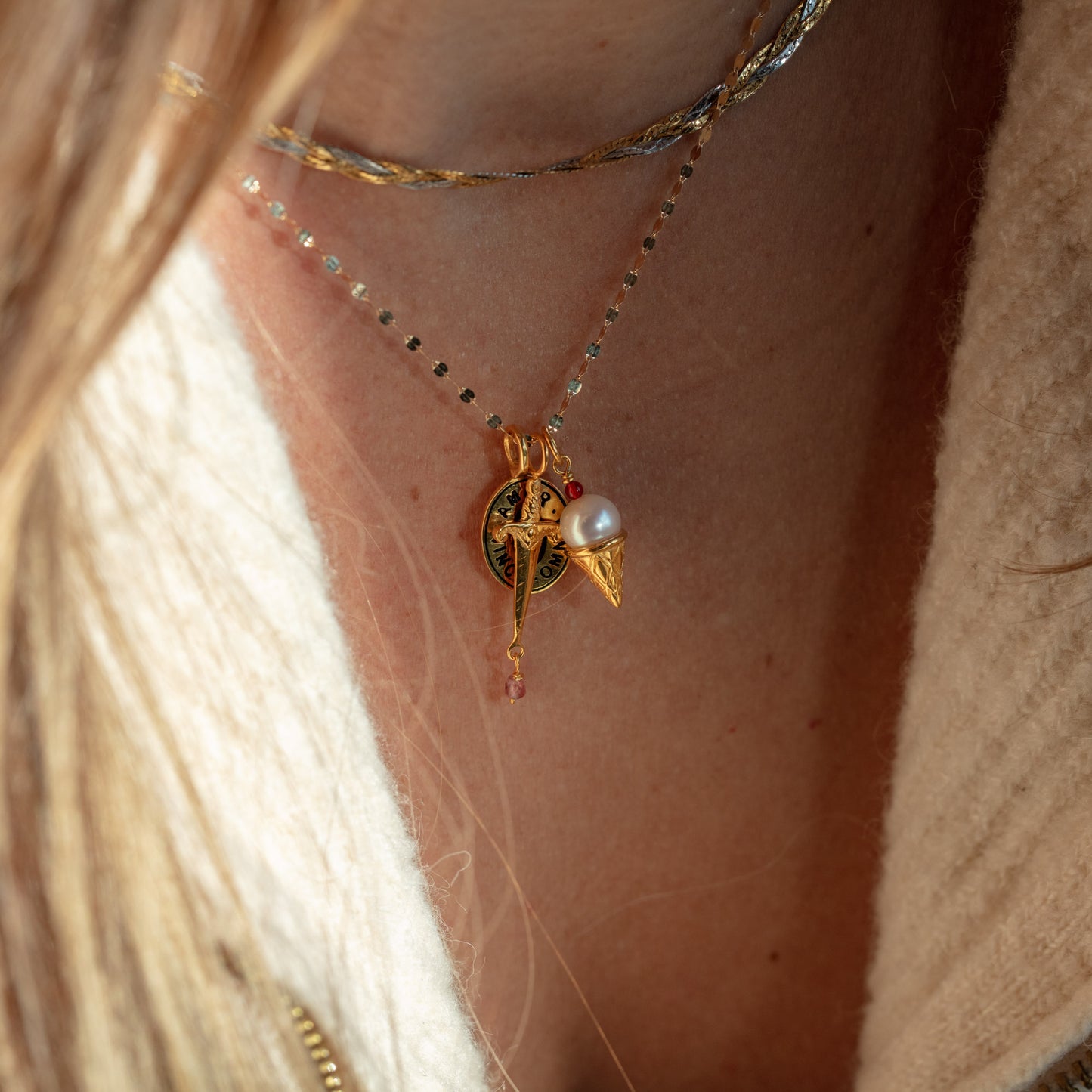 Close-up of a person wearing three layered Charms by Hunt of Hounds necklaces: a thin sterling silver chain, one with a gold vermeil pendant, and another with gold, sword, and white pearl charms over a cream-colored top.