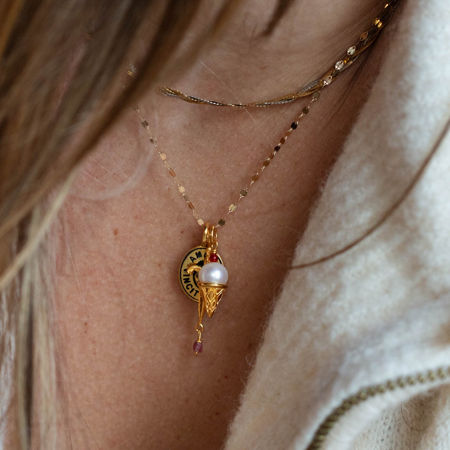 Close-up of a persons neck wearing three Charms by Hunt of Hounds gold vermeil necklaces: one with a LUCKY round charm, one with a pearl in a gold setting, and one with a small pink gemstone. The person wears a cream-colored top.