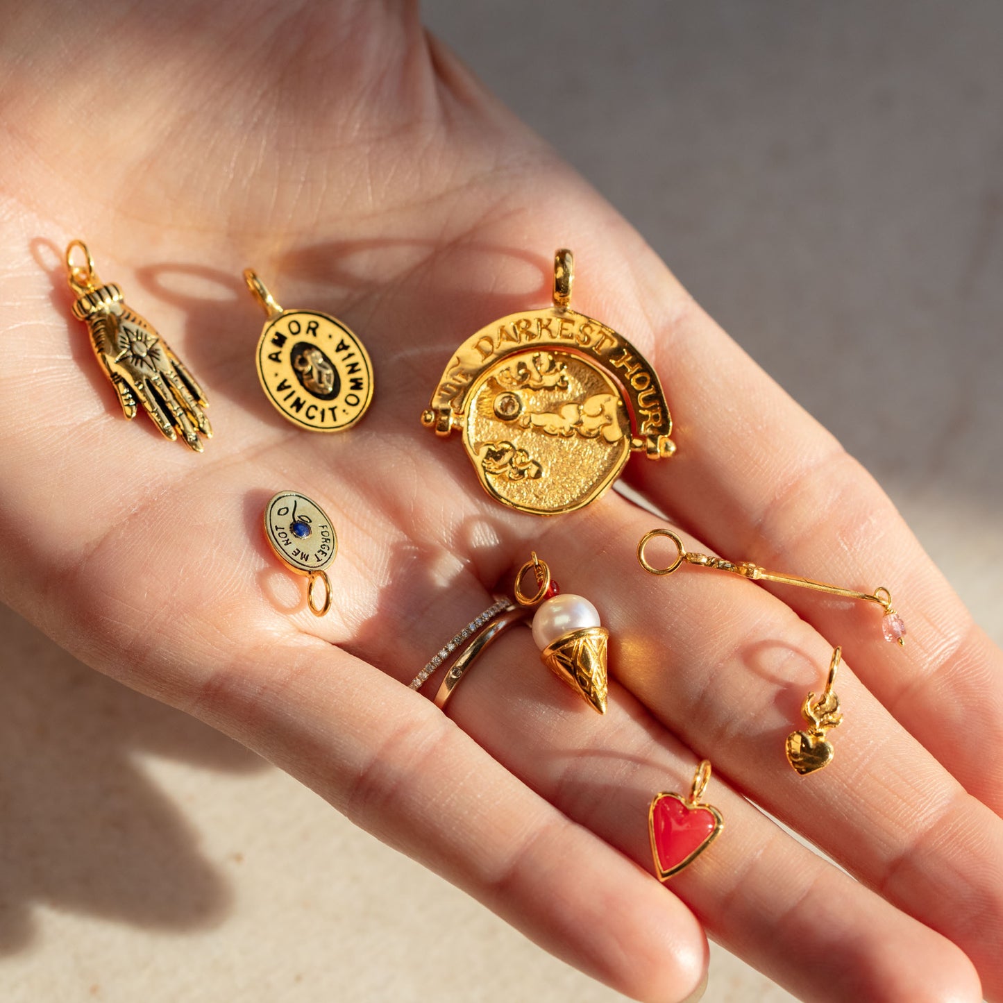 A hand displays Charms by Hunt of Hounds, featuring a sun and moon hand, Viva or Vino Vinci circle, In My Darkest Hour arc, pearl ice cream cone, red heart, gold heart charms, and more.