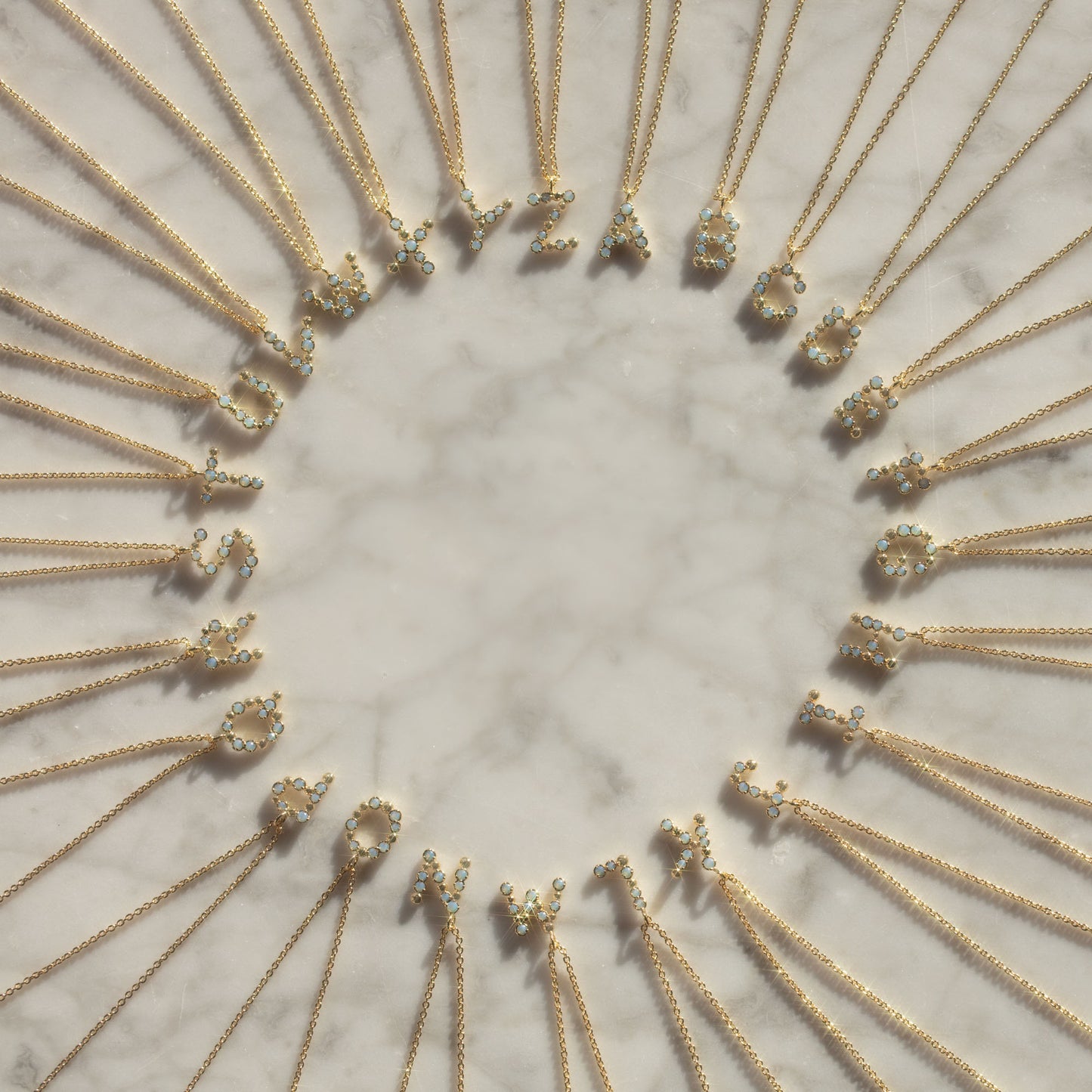 Initial Charm Necklace Sample features brass-based, 14k yellow gold-plated necklaces with opalescent nano gem letter pendants, arranged in a circle on marble and evenly spaced.