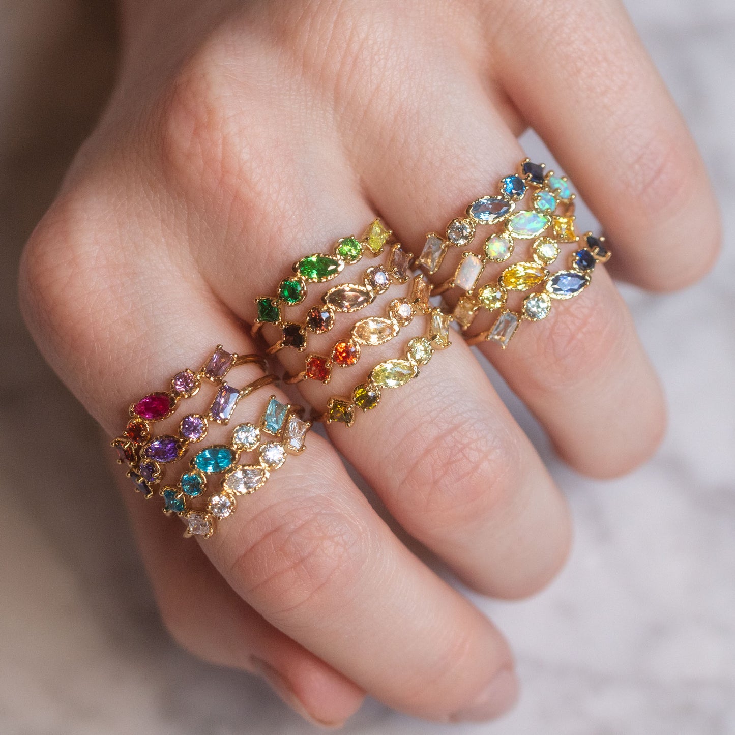 A hand showcases the Shades of You Ombre Birthstone Ring, 14k gold plated and set with colorful birthstones in various shapes and sizes, against a softly blurred background.
