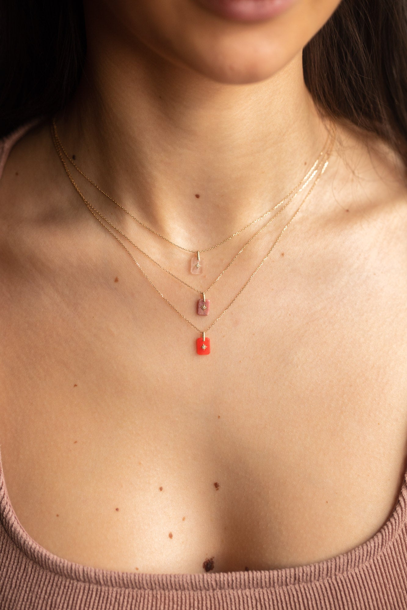 A woman wears three Solid Gold Crystal Pendant Necklaces—clear, rose quartz pink, and red—layered over a sleeveless ribbed pink top. The image is cropped to her neck and upper chest.