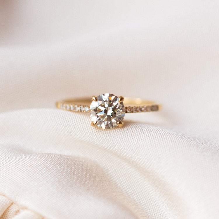 The 9kt Round Cut Diamond Solitaire Engagement Ring with Pave Diamond Band (Sample Size 7) is displayed on a soft, cream-colored fabric background.