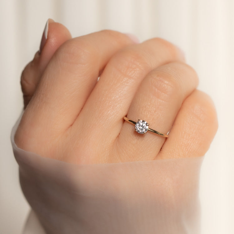 A hand with a sheer sleeve showcases the 9k Classic Round Cut Diamond Solitaire Engagement Ring Sample Size 7, featuring a center round lab created diamond on a yellow gold band, set against a softly blurred light background.