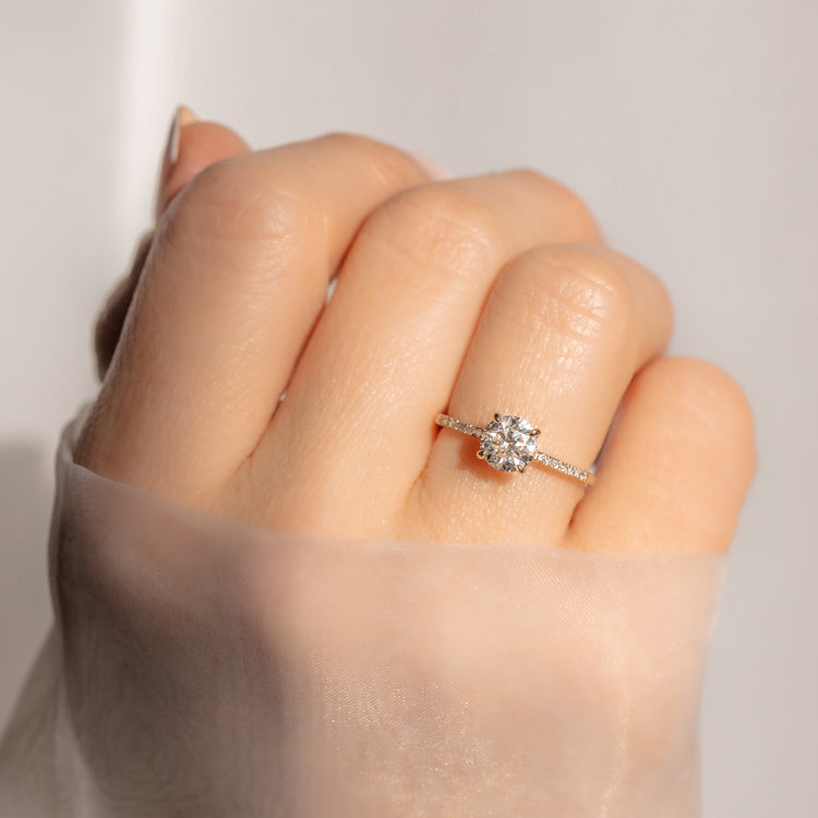 A hand draped in sheer fabric wears the 9kt Round Cut Diamond Solitaire Engagement Ring with Pave Diamond Band, sample size 7—a sparkling lab-created diamond set on a slim yellow gold band encrusted with stones.