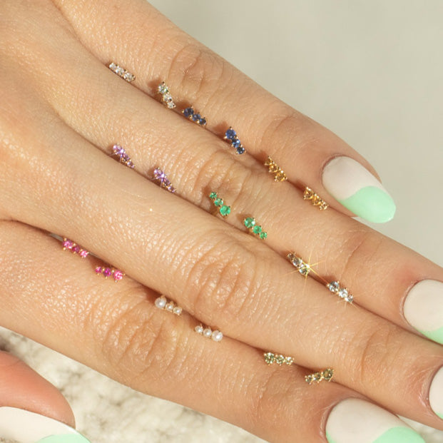 A close-up of hands with pastel green and white manicured nails, each finger adorned with colorful gemstone rings and Solid Gold Dainty Birthstone Bar Studs earrings, set against a soft, neutral background.