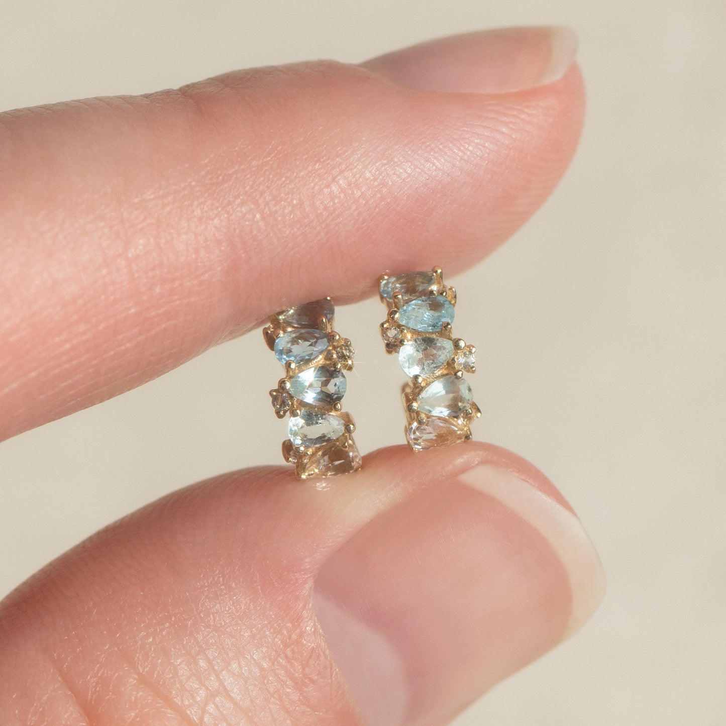 A hand holds Solid Gold Ombre Birthstone Huggie Hoops featuring 9k gold, three light blue gemstones, and clear stones against a neutral background—elegant birthstone earrings for any occasion.