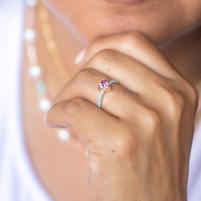 A medium brown-skinned person in a white top and layered necklaces rests their chin on their hand, showcasing the Little Love Ring—plated in 14k yellow gold with a pink gemstone—on their finger.