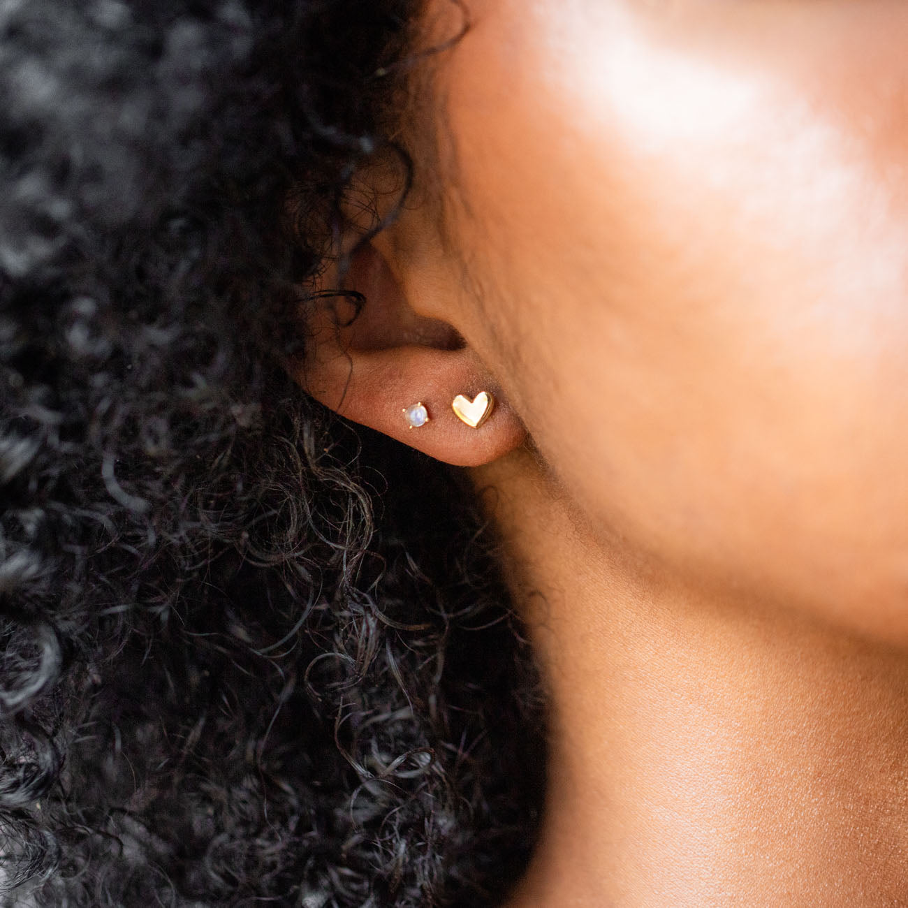 Close-up of a person’s ear with two earrings: a small round stud and the Solid Gold Moonstone Studs. The person has curly black hair, warm brown skin, and an elegant, understated style.