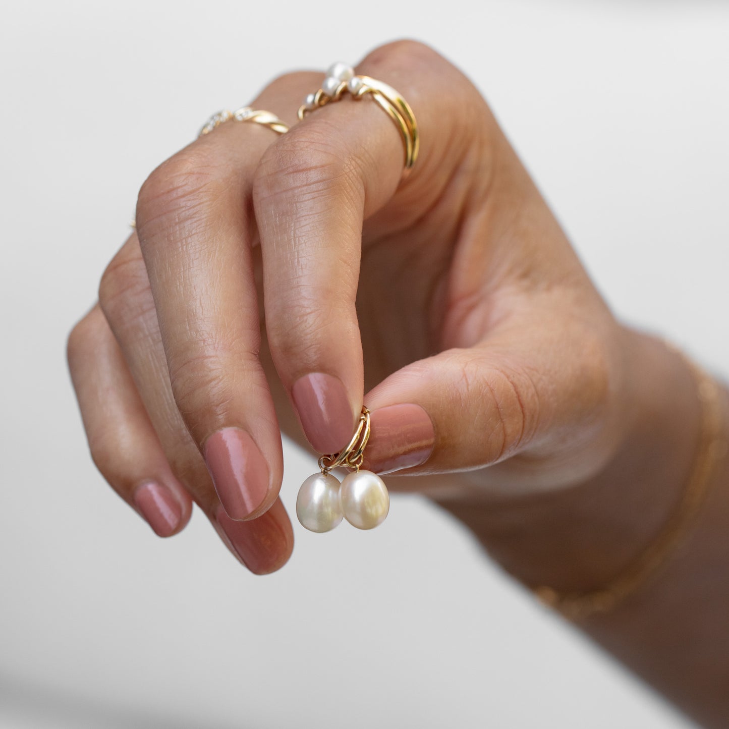 A hand with pink nail polish holds the Solid Gold Elegant Pearl Charm Huggies, surrounded by gold and pearl rings, against a soft, blurred background.