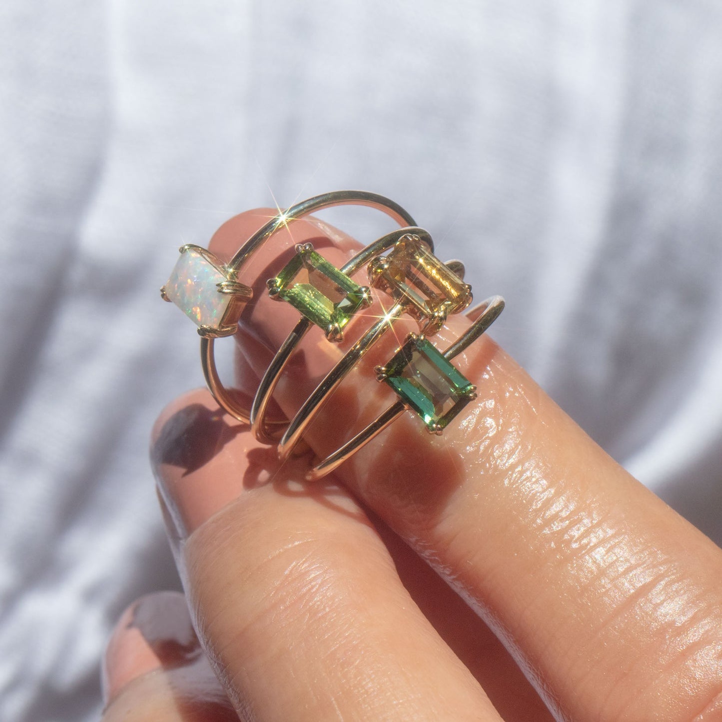 A close-up of a hand holding the Solid Gold Essential Baguette Birthstone Ring in Blue Sapphire, featuring three yellow gold bands with sparkling rectangular gemstones, including blue sapphire, gleaming brightly in the sunlight.