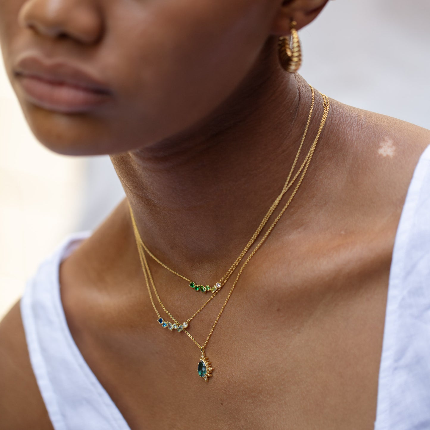 A woman in a white top is shown from the shoulders up, layering gold necklaces and hoops, with focus on her Shades of You Ombre Birthstone Necklace featuring green and blue gemstone pendants for a personalized touch.