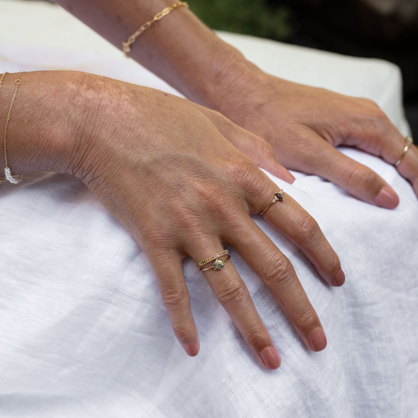 Two hands with manicured nails rest on a white cloth, adorned with delicate gold bracelets and the Solid Gold Live by the Sun Rainbow Ring. Soft lighting enhances the rings subtle sparkle.