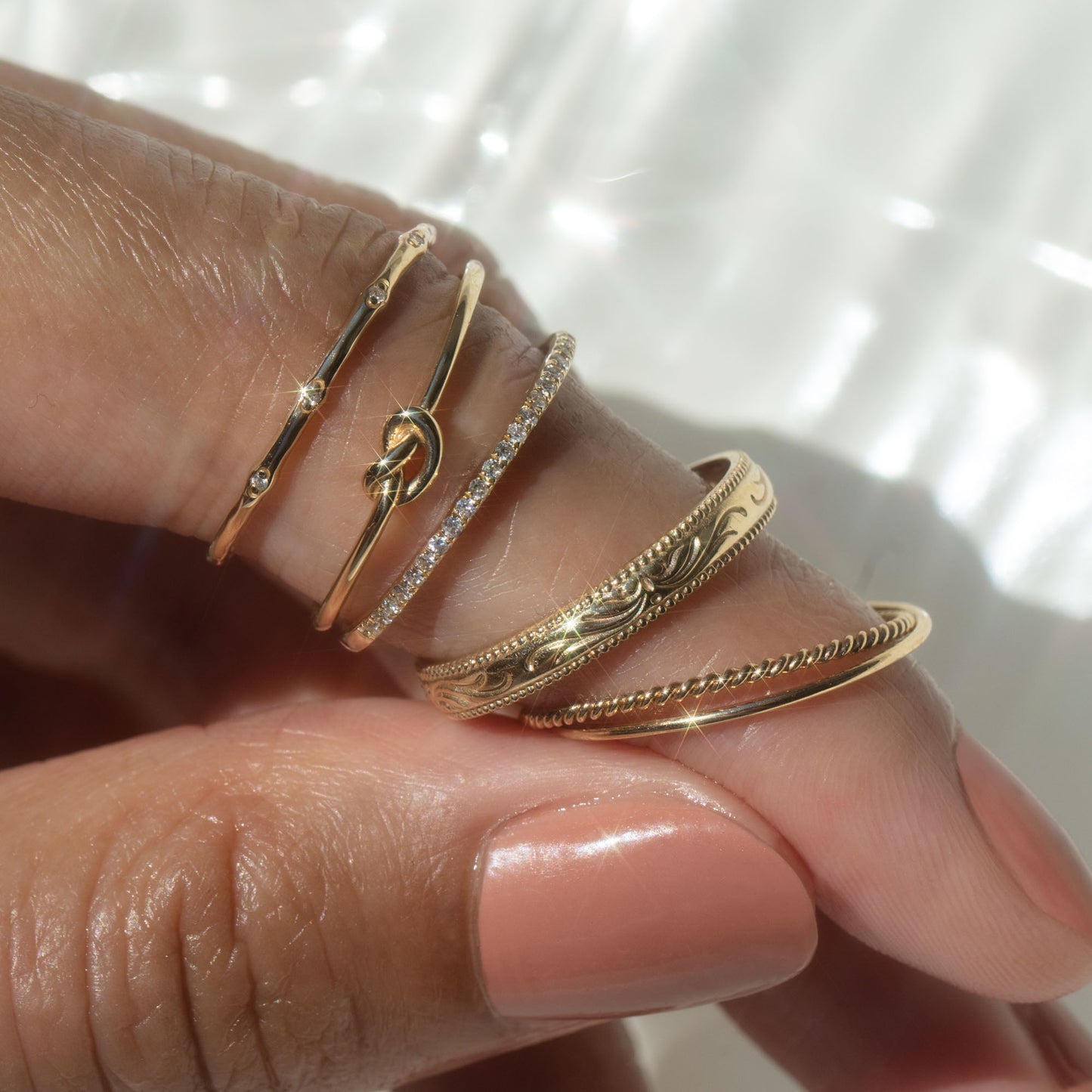 A hand with glossy nude-pink nails wears four delicate rings, including the Solid Gold Simple Knot Ring in 10k yellow gold, along with styles featuring small diamonds, engraved patterns, and a twisted detail against a softly lit background.