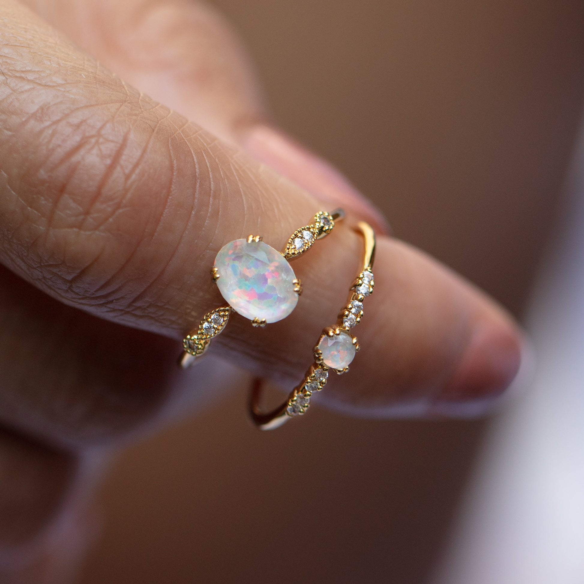 A close-up of a hand wearing two Opal Sparkle Rings, each crafted in gold with shimmering opals and small clear gemstones, set against a softly blurred background.