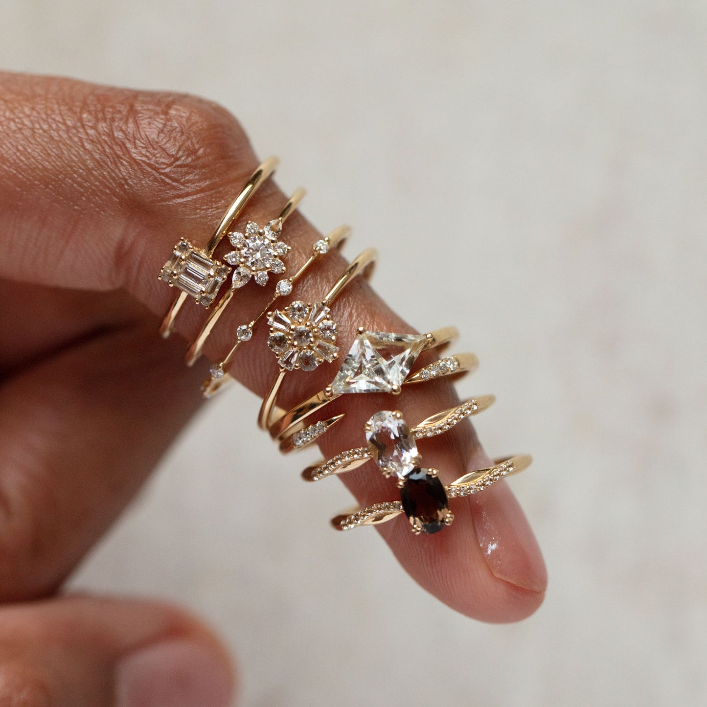 A hand with four fingers, each wearing stacked gold rings made from recycled gold and gemstone shapes—round, square, oval—and a 14k Floral Art Deco Ring with white sapphire accents, all set against a neutral background.