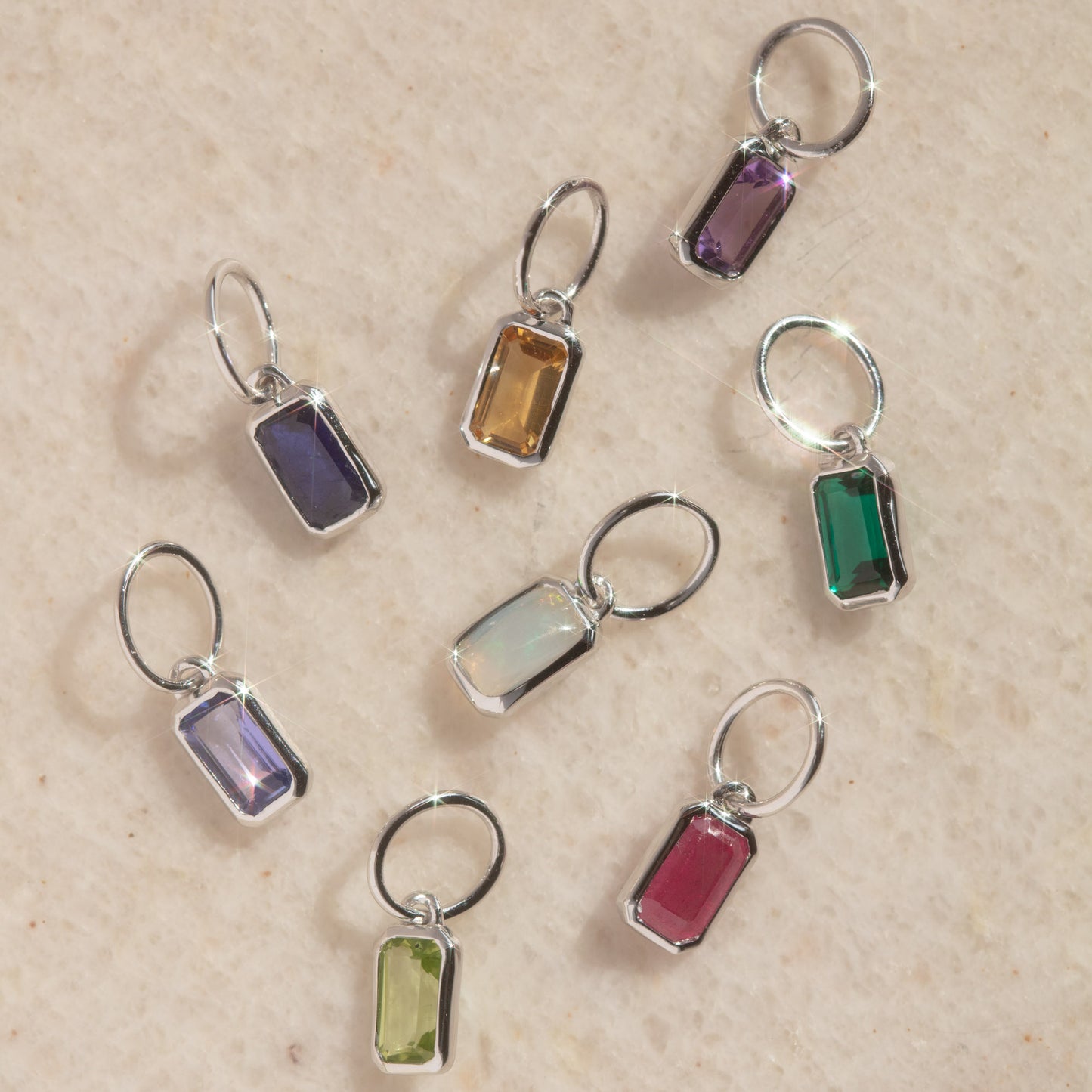 The Solid Gold Birthstone Baguette Charm in White Gold features eight rectangular gemstone charms, each a unique color set in white gold, arranged on a light beige textured surface for an elegant, personalized touch.