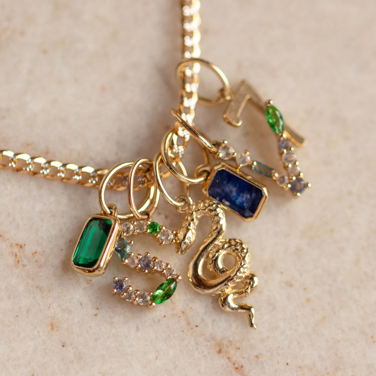 The Solid Gold Gemstone Letter Charm necklace features several pendants, such as a personalized gold charm, a gold snake, green and blue rectangular gemstones, a gold key, and sparkling stones displayed on a beige surface.