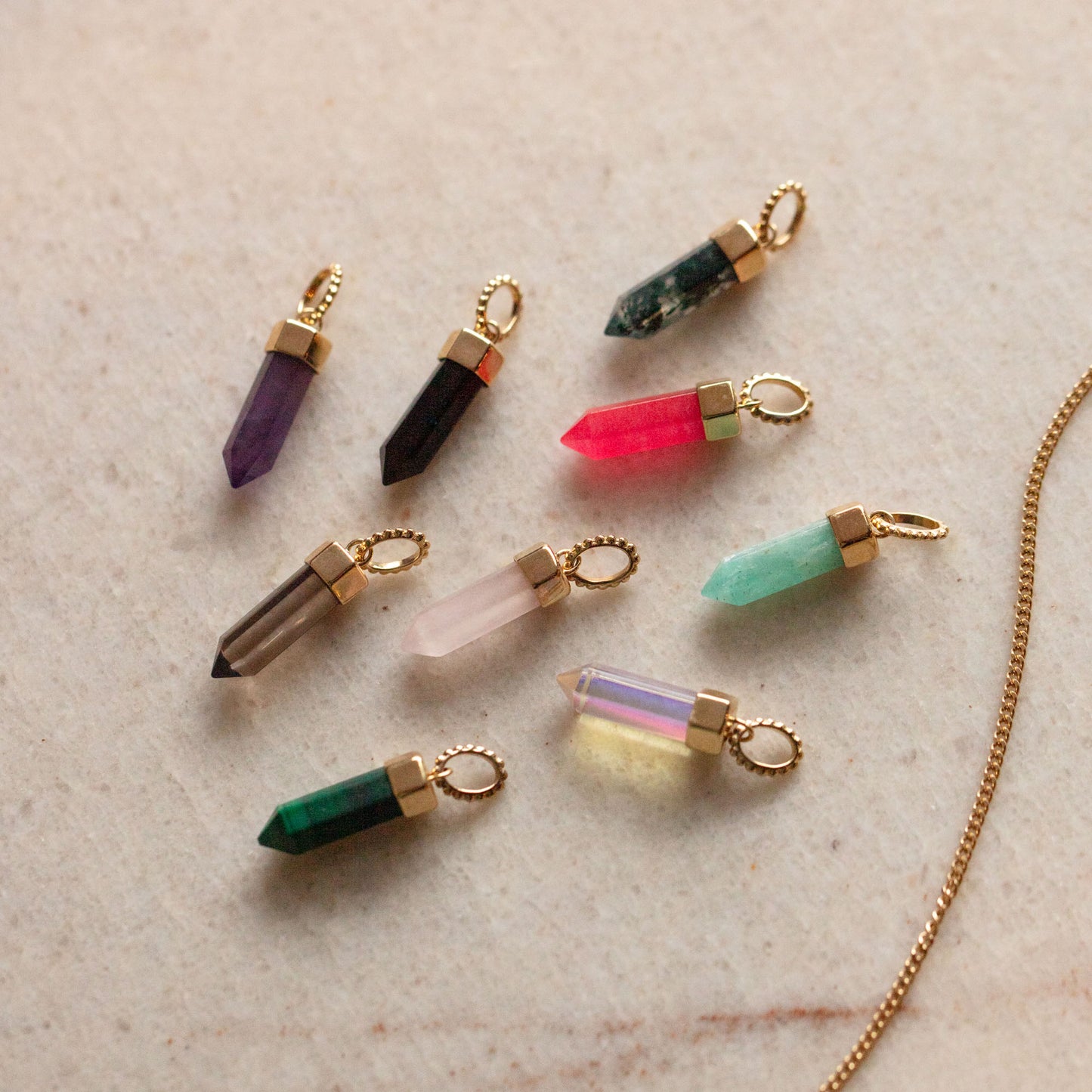 Nine Solid Gold Crystal Charms for Manifestation, each featuring a unique color—purple, pink, green, black, and clear—are displayed on a light textured background. These natural crystal pendants come with gold loops for added style.