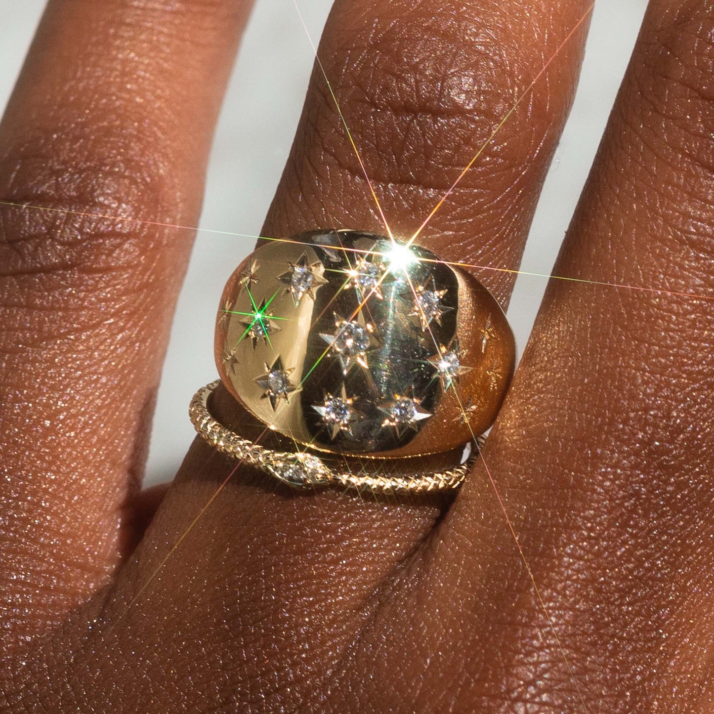 A close-up of a hand wearing two rings: the Ouroboros Diamond Snake Ring in shiny 14k solid gold with star-shaped diamonds, and a twisted gold band below, both glinting in bright light that creates a lens flare.