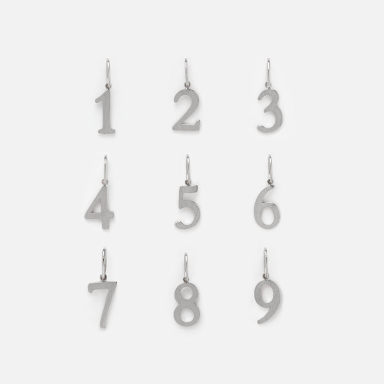 Nine solid white gold number charms arranged in three rows. Numbers 1 to 9 each have a small loop for attaching to a chain or bracelet—ideal for personalized jewelry. The background is plain white. Product: Solid Gold Number Charm in White Gold.