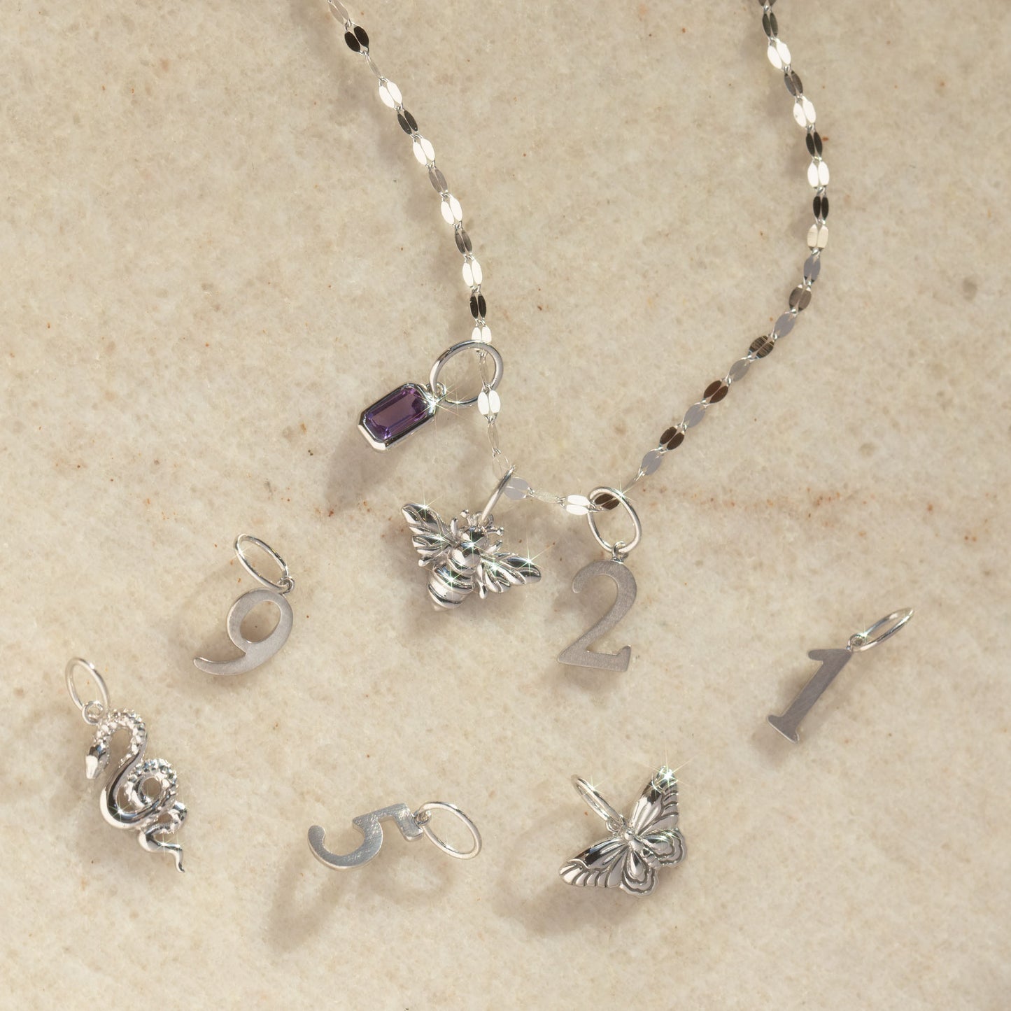 A personalized jewelry piece features a silver chain with a purple gem, bee charm, and a Solid Gold Number Charm in White Gold (2), displayed on beige, alongside loose charms: dragon, numbers 9 & 5 in gold & number 1.