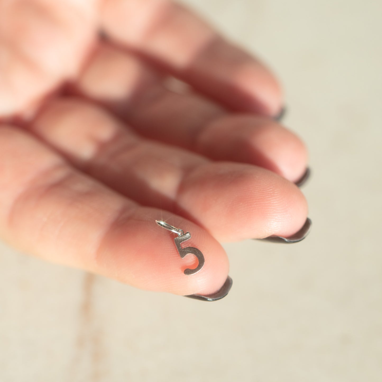 A hand with black manicured nails holds the Solid Gold Number Charm in White Gold, shaped like the number 5, against a light background.