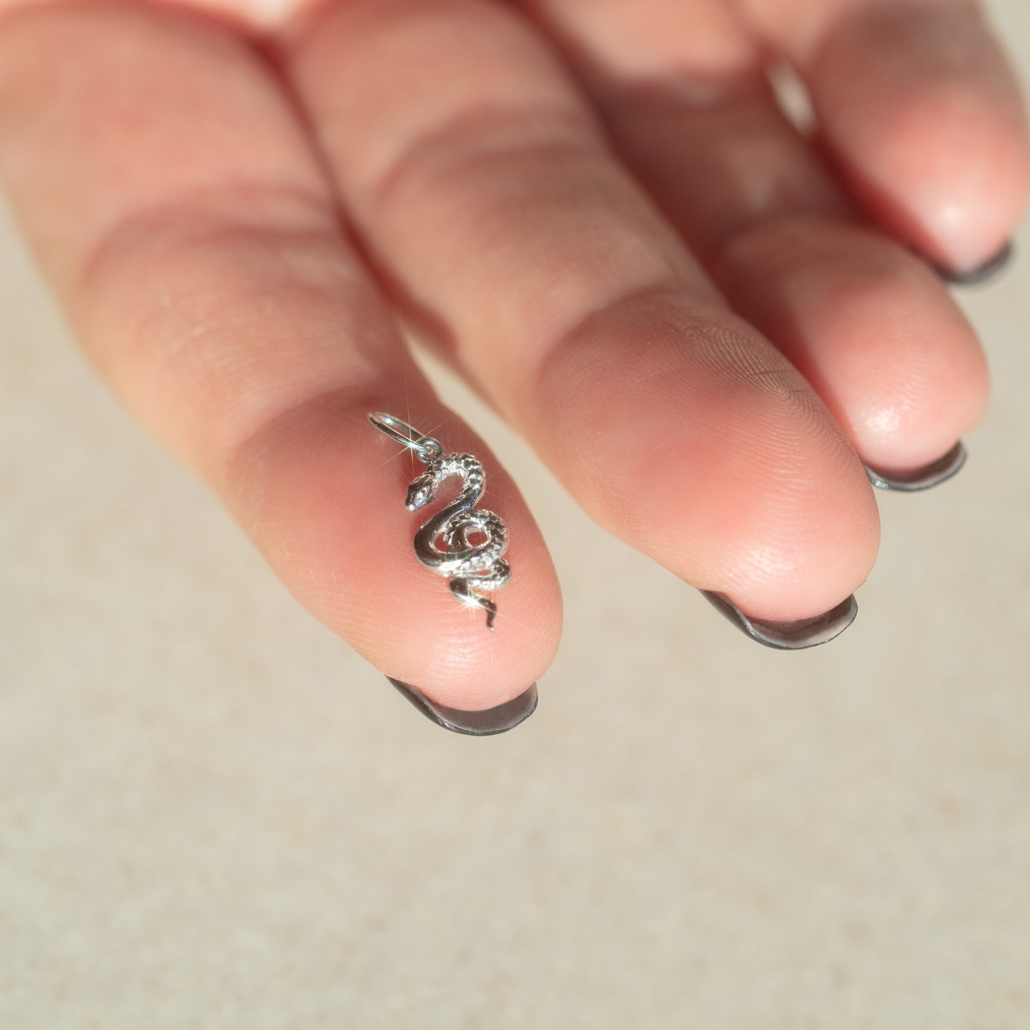 A close-up of a hand with black-tipped nails holds the Solid Gold Nature Charm in White Gold—a small, snake-shaped pendant with delicate pearl detail—between two fingers, against a softly blurred background.