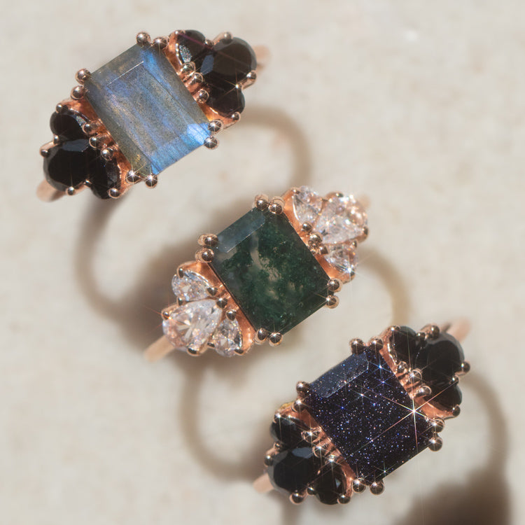 Three Gemstone Empress Rings—gold with blue, green, and dark blue rectangular gemstone centers, accented by clear and black stones—are displayed on a lightly textured surface.