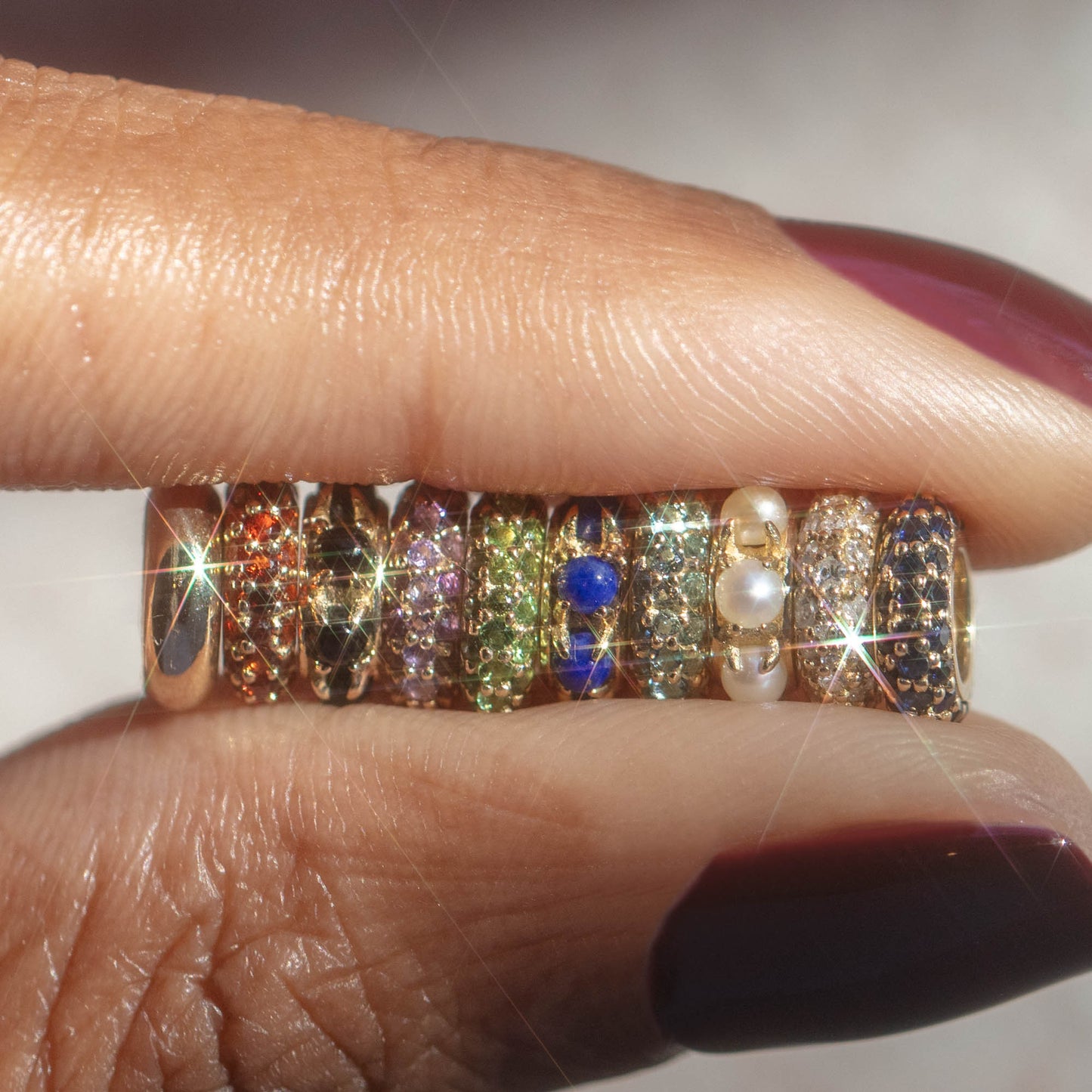 A hand with burgundy nail polish holds nine sparkling, colorful gemstone rings and a pearl-accented ring, showcasing the Solid Gold Anniversaire Charm in bright light that highlights each milestone-inspired jewelry piece.