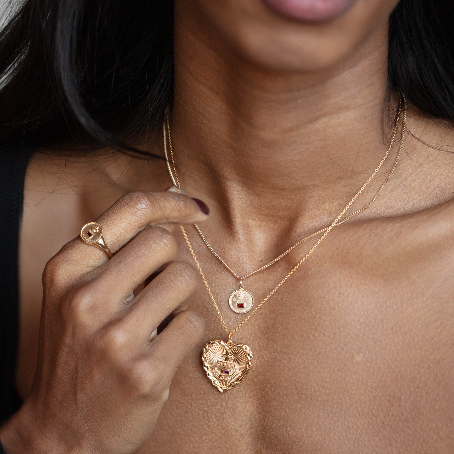 A close-up of a woman wearing layered gold necklaces, featuring the Solid Gold Coin Charm and a heart-shaped locket, along with a gold ring on her finger as her hand rests near her collarbone.