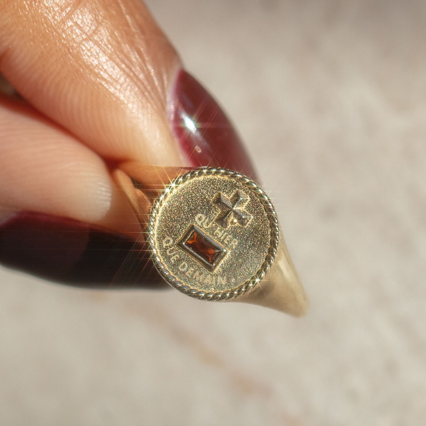A hand with long, polished nails holds the Solid Gold Quhier Que Demain Signet Ring, its engraved circular face sparkling brightly against a blurred background.
