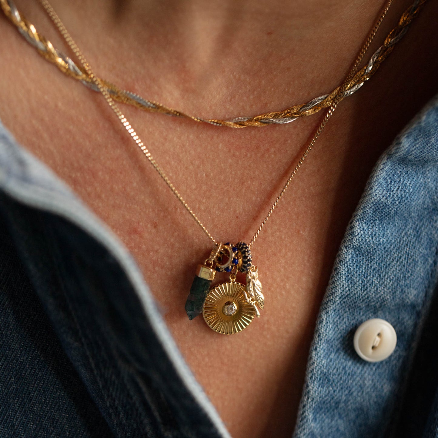 Close-up of a person’s neck wearing layered necklaces, featuring the Solid Gold Crystal Charm for Manifestation among other gold chains and gemstone pendants, paired with an open denim shirt and white button.