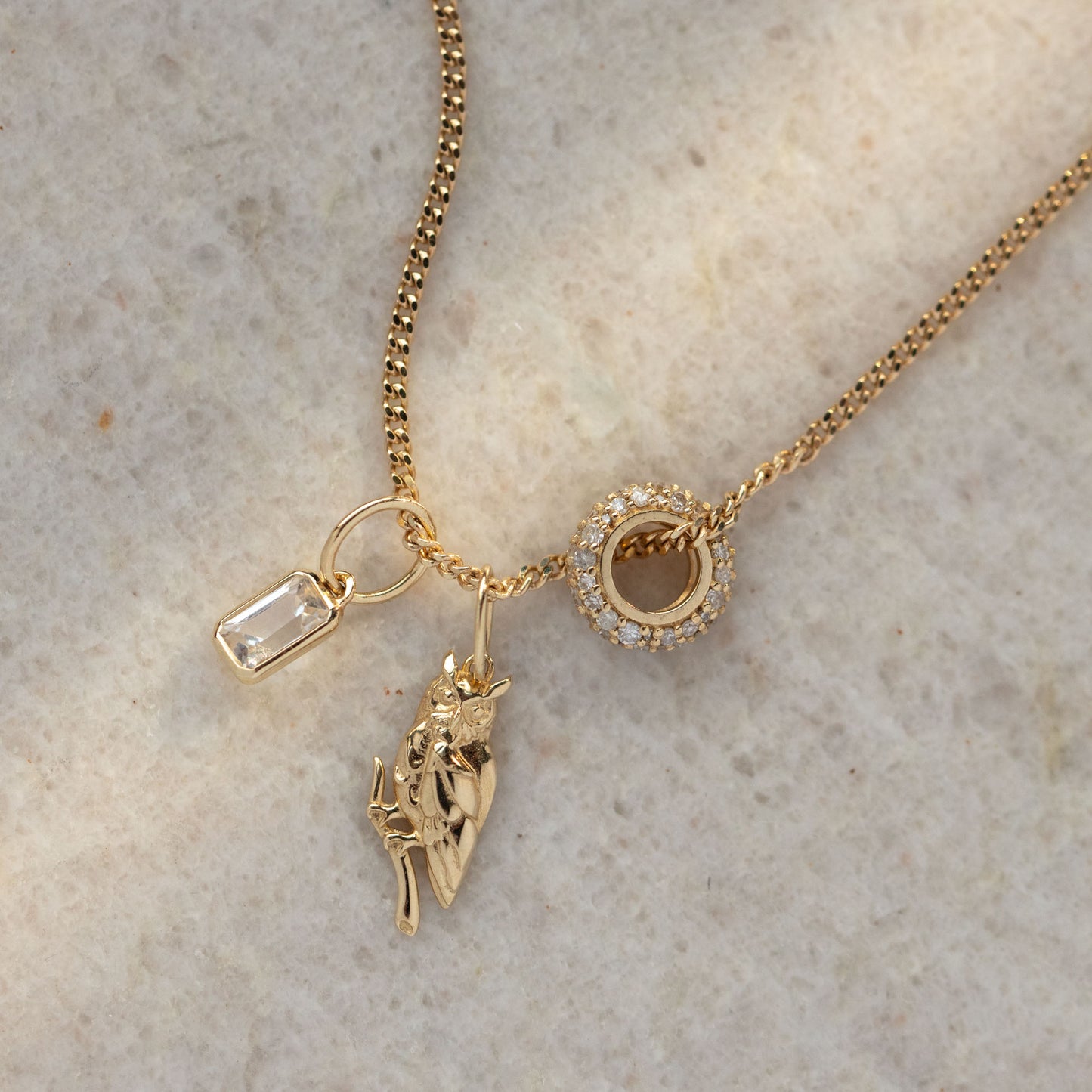 The Solid Gold Anniversaire Charm necklace showcases trois charms: a clear rectangular gemstone, an anatomical heart, and a sparkling circular pendant—ideal milestone jewelry, elegantly set against a light stone background.