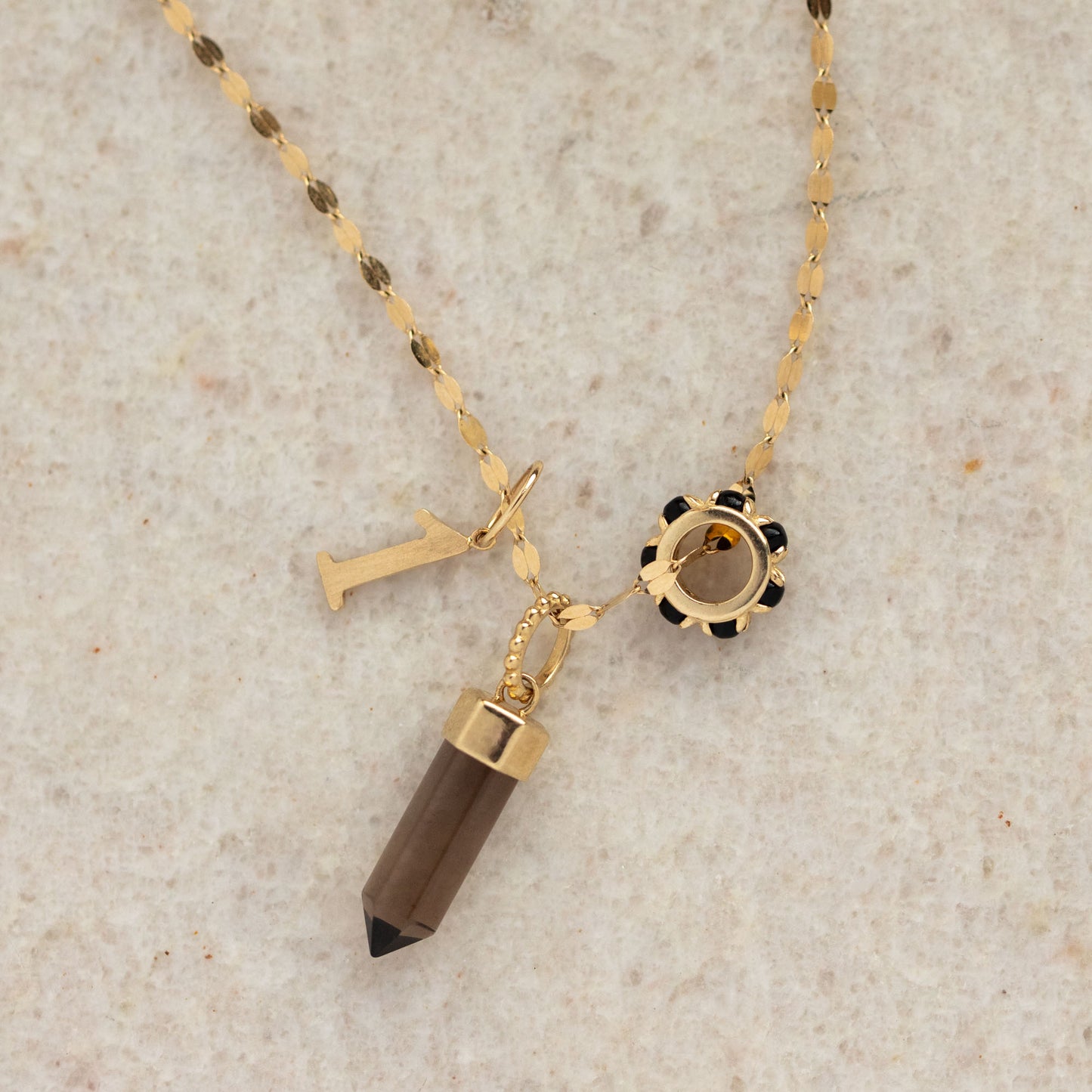 A Solid Gold Anniversaire Charm necklace with three charms—a vertical letter I, a round black and gold floral charm, and a brown crystal pendant—perfect for milestones or celebrations, displayed on a light textured surface.