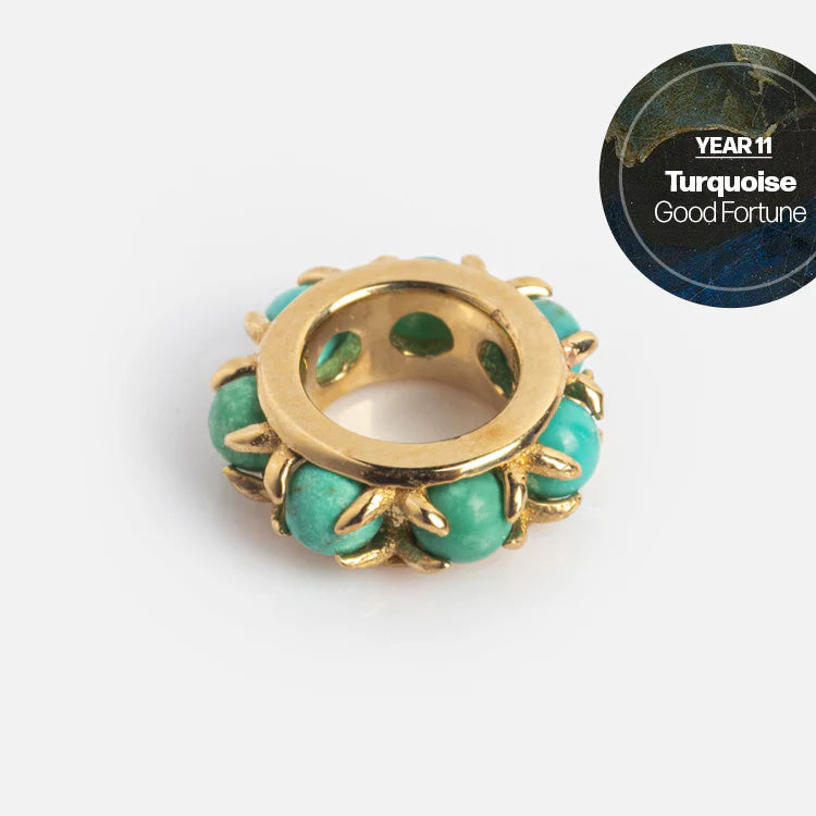 A gold circular Solid Gold Anniversaire Charm with turquoise stones on a white background. In the top right, a dark circle says YEAR 11, Turquoise, Good Fortune—celebrating milestone jewelry moments.