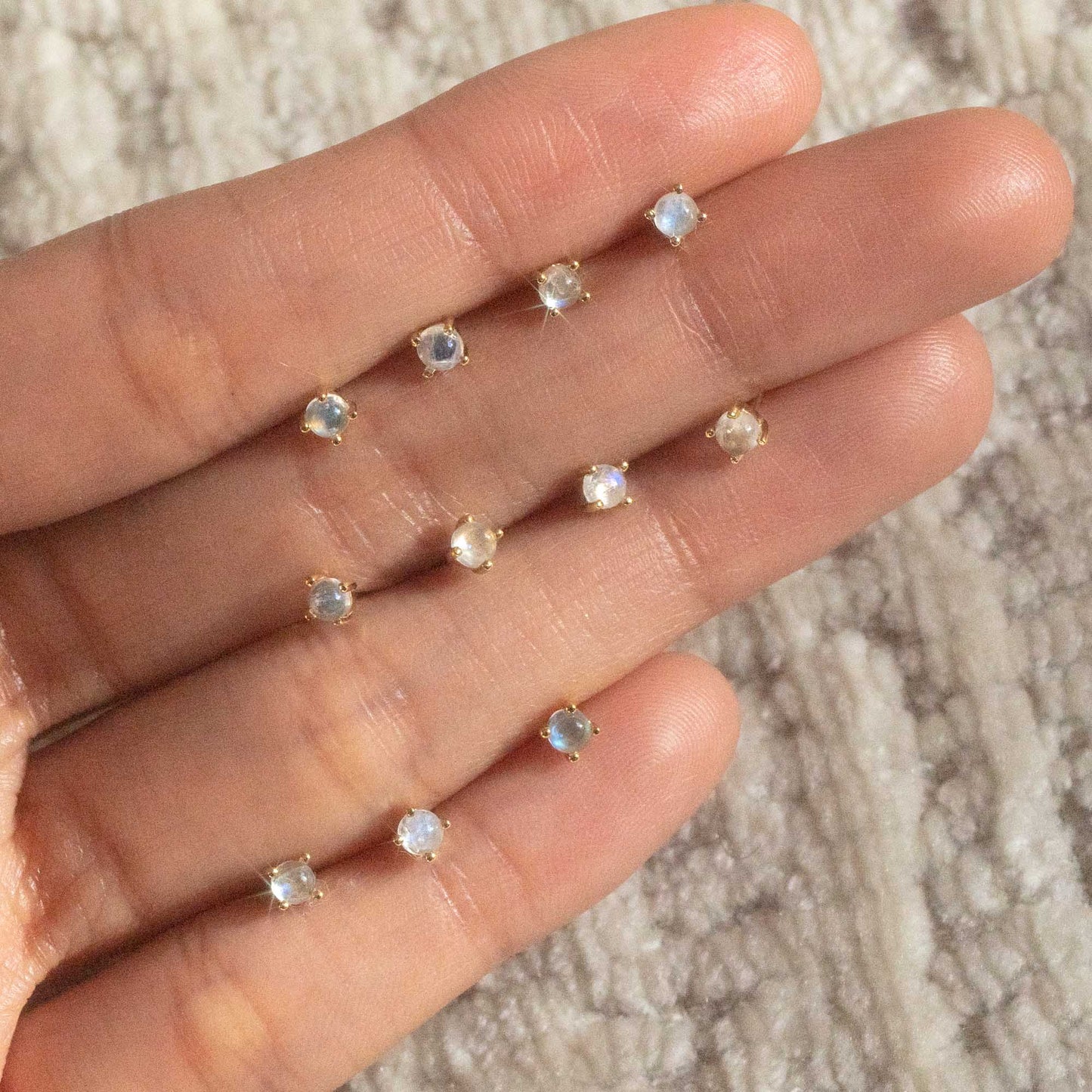 A hand displays ten Solid Gold Moonstone Studs, each featuring a translucent moonstone set in solid gold, arranged in a row across the fingers on a textured background.