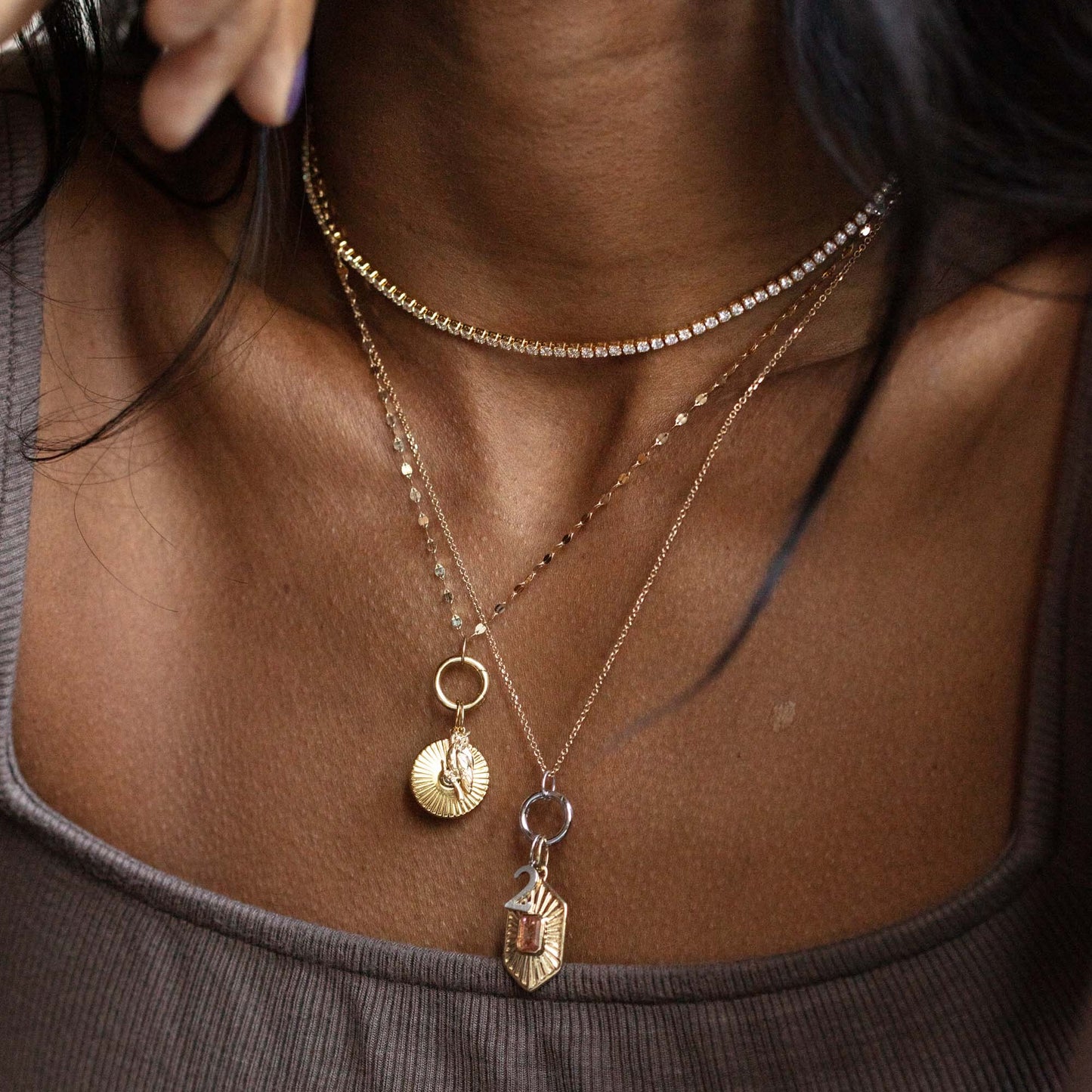 A close-up of a person wearing three layered necklaces, including the Solid Gold Charm Clip in White Gold, over a ribbed taupe tank top. Their dark hair partially frames their neck and shoulders.