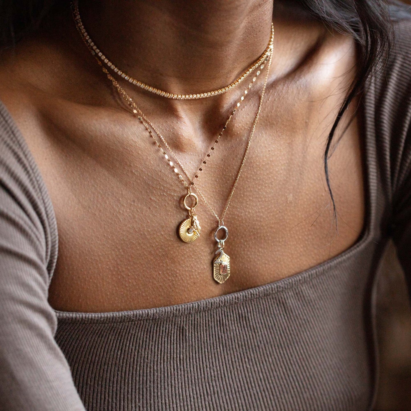A person in a ribbed taupe off-the-shoulder top showcases three layered gold necklaces, featuring the Solid Gold Amulet Charm. Warm-toned skin and loose hair frame their neck and shoulders.