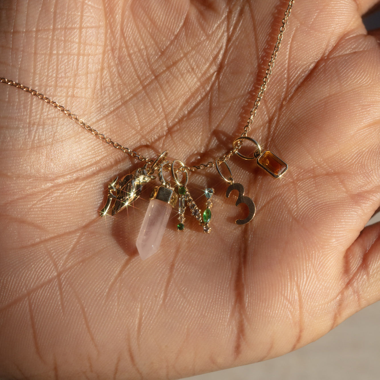 A hand holds the Solid Gold Gemstone Letter Charm necklace, featuring a gemstone letter, pink crystal, small gemstones, a star, the number 3, and an amber gem—all sparkling in sunlight.