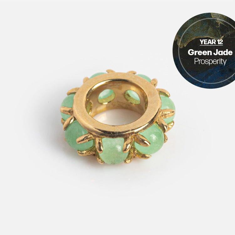 The Solid Gold Anniversaire Charm is a circular gold bead set with green jade stones, symbolizing prosperity. The jewelry features “YEAR 12 Green Jade Prosperity” in a dark circle at the top right corner.