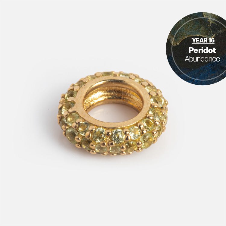 The Solid Gold Anniversaire Charm, a ring-shaped bead adorned with small peridot stones, shines brightly. In the top right corner, “YEAR 16 Peridot Abundance” appears on a dark, marble-like background.