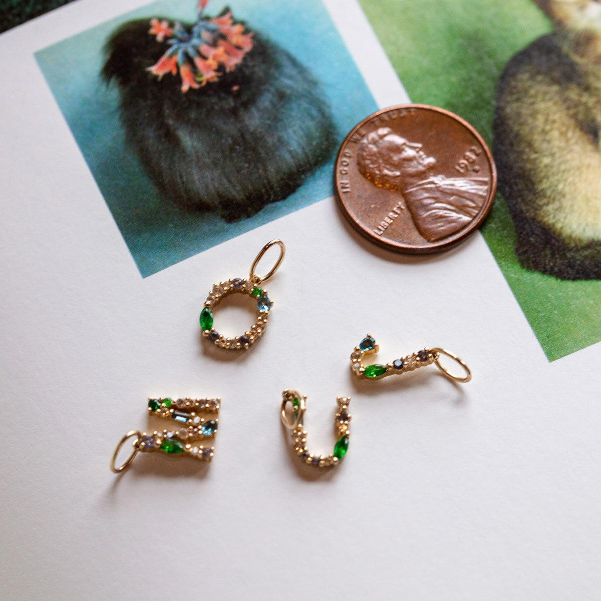 Four personalized Solid Gold Gemstone Letter Charms with green and clear stones are arranged on paper near a U.S. penny for scale, positioned next to images of furry animals.