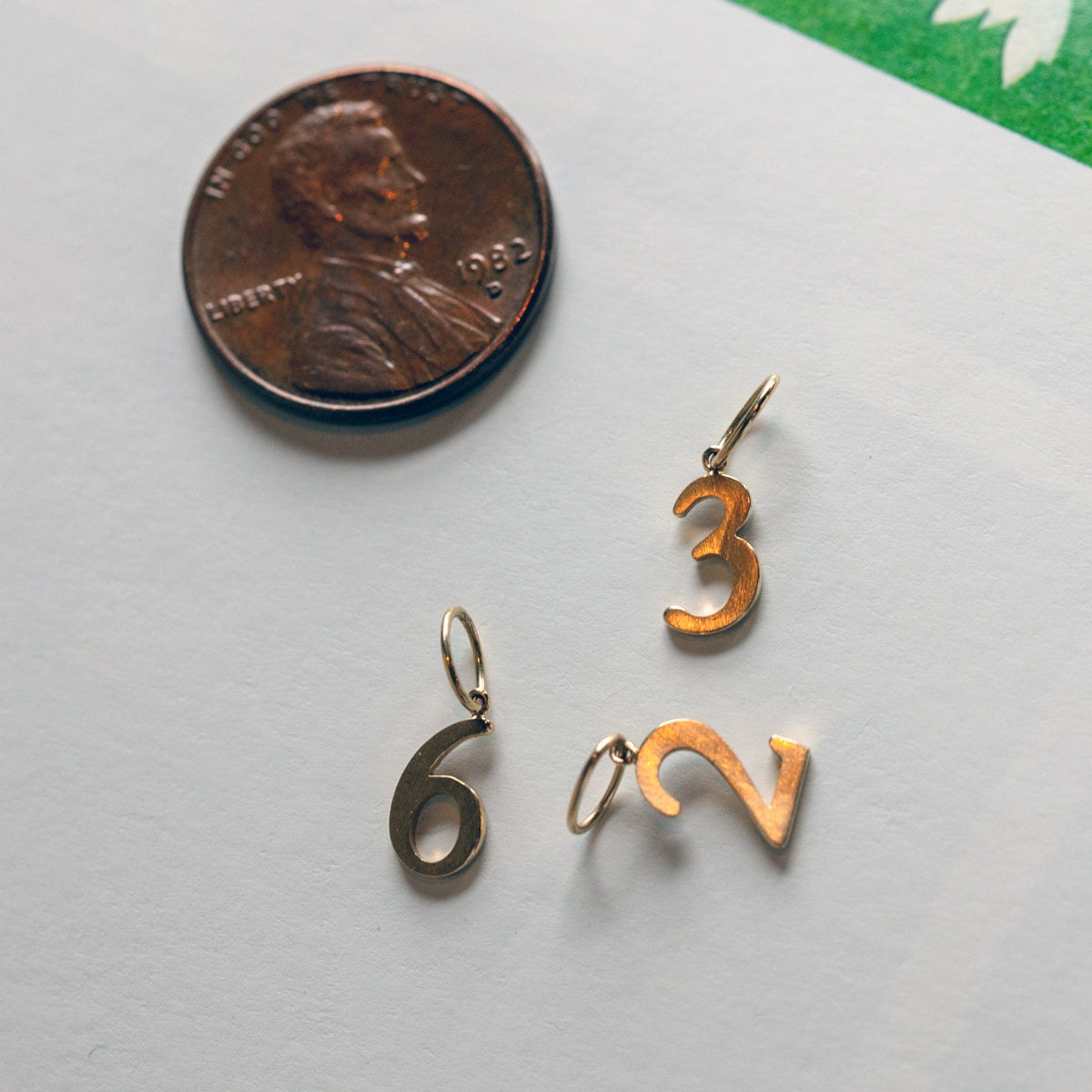 A 1982 US penny is next to three Solid Gold Number Charms—6, 3, and 2—on a white surface with a green corner showing at the top right, ideal for personalized jewelry lovers.