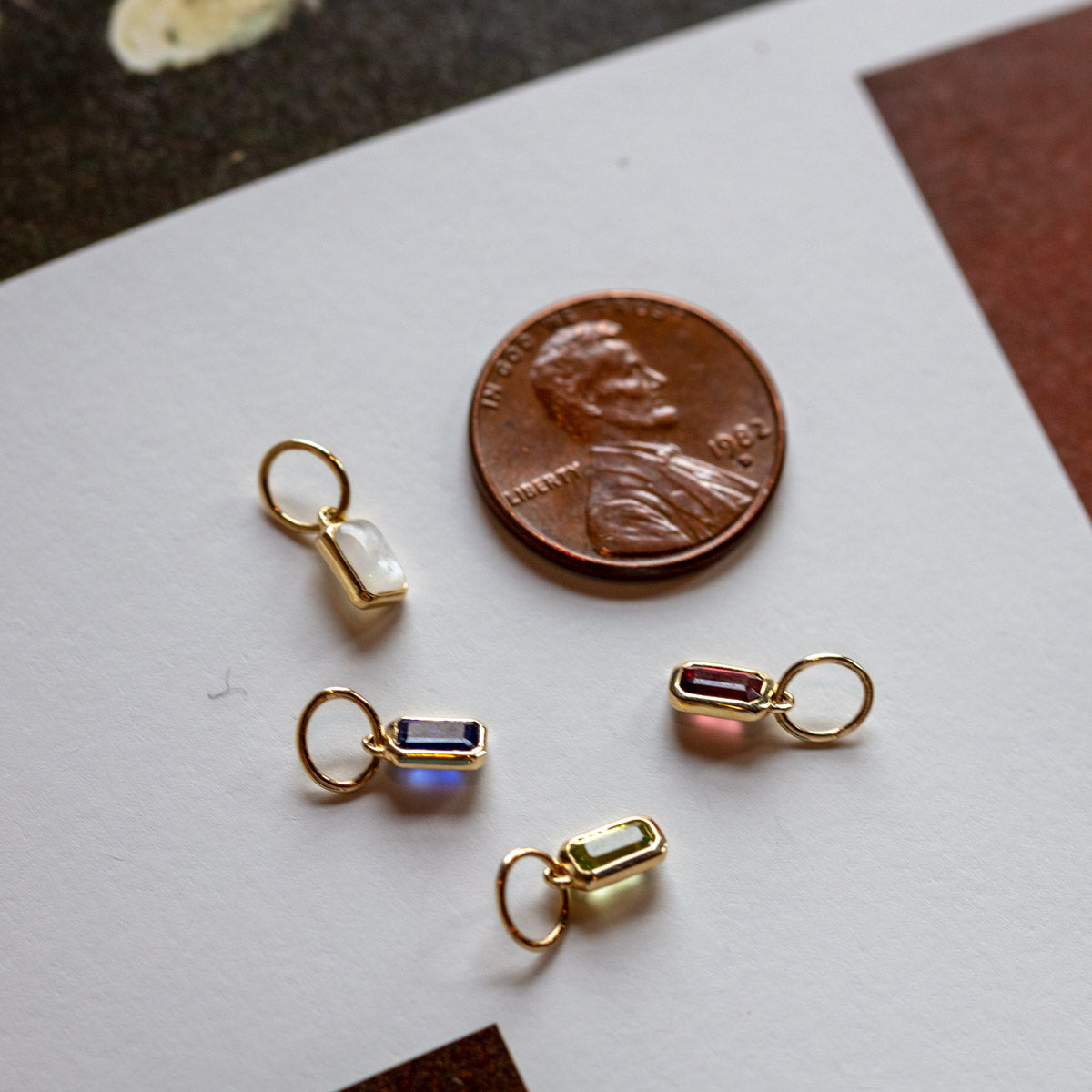 A U.S. penny next to four Solid Gold Birthstone Baguette Charms in white, blue, pink, and light green on a white surface. These charms are ideal for personalized jewelry and are all smaller than the penny.