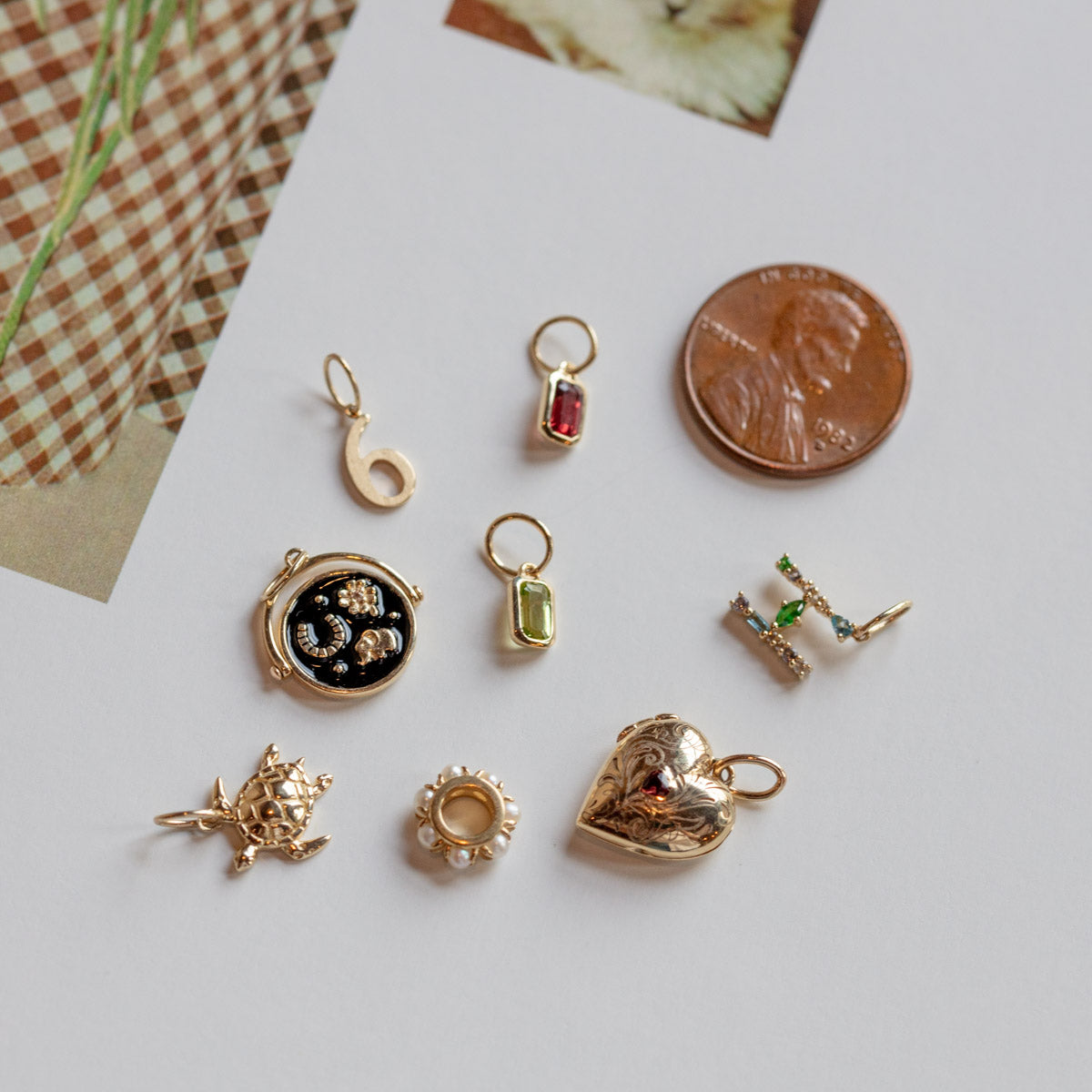 A collection of Solid Gold Anniversaire Charm designs—including a heart, black circular charm, hook, and flower—shown on paper beside a US penny for scale; ideal as anniversary or milestone jewelry accents.