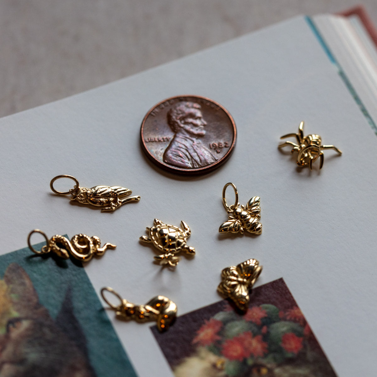 A penny and seven Solid Gold Nature Charms—including a cat, turtle, bee, spider, dragon, butterfly, and frog—are displayed on an open book with colorful illustrations, highlighting the whimsical charm of spiritual jewelry.