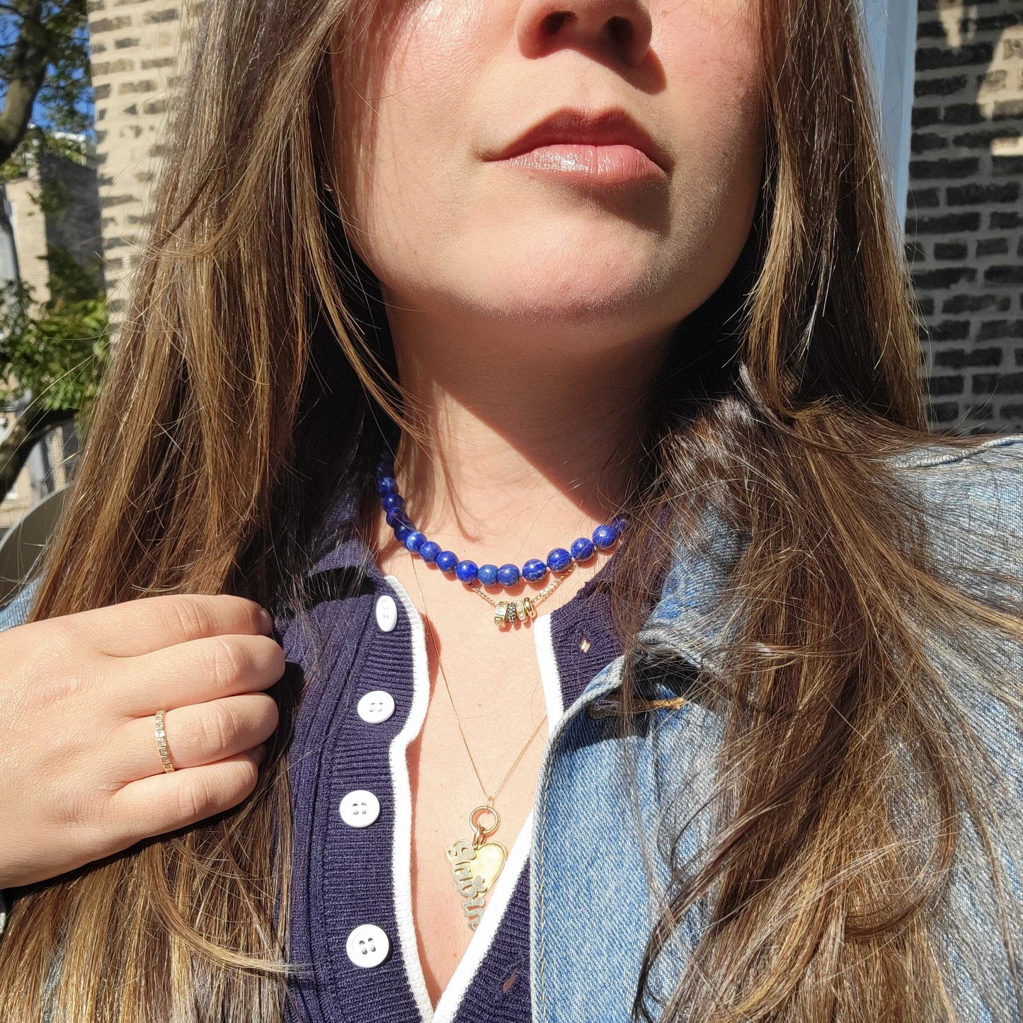 A woman with long brown hair wears the Sale! Beaded Gemstone Necklace, paired with a navy collared top and denim jacket, standing outside in sunlight against a brick wall.