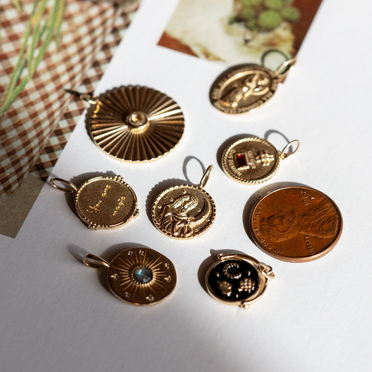 A set of eight Solid Gold Coin Charms featuring various embossed designs and small gemstones, displayed on a white surface with patterned and photo papers in the background.